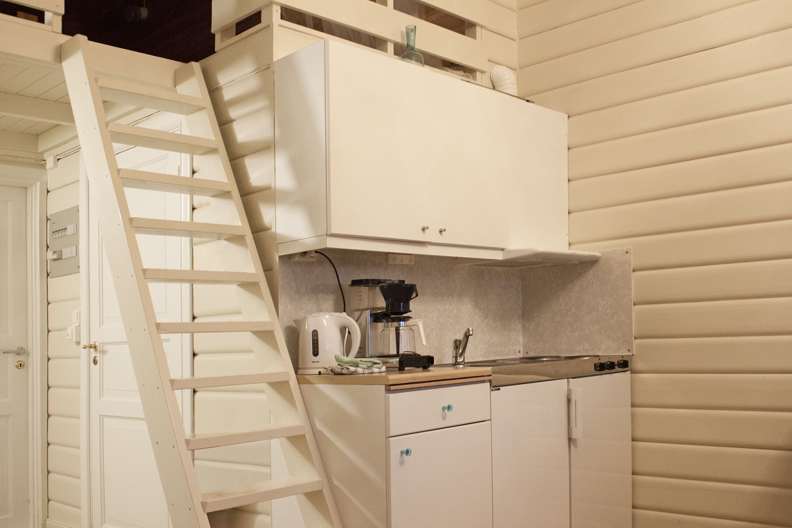 Cocina compacta con estufa integrada, cafetera y lavabo. Una escalera de madera está apoyada contra la pared, dando acceso a un nivel superior.
