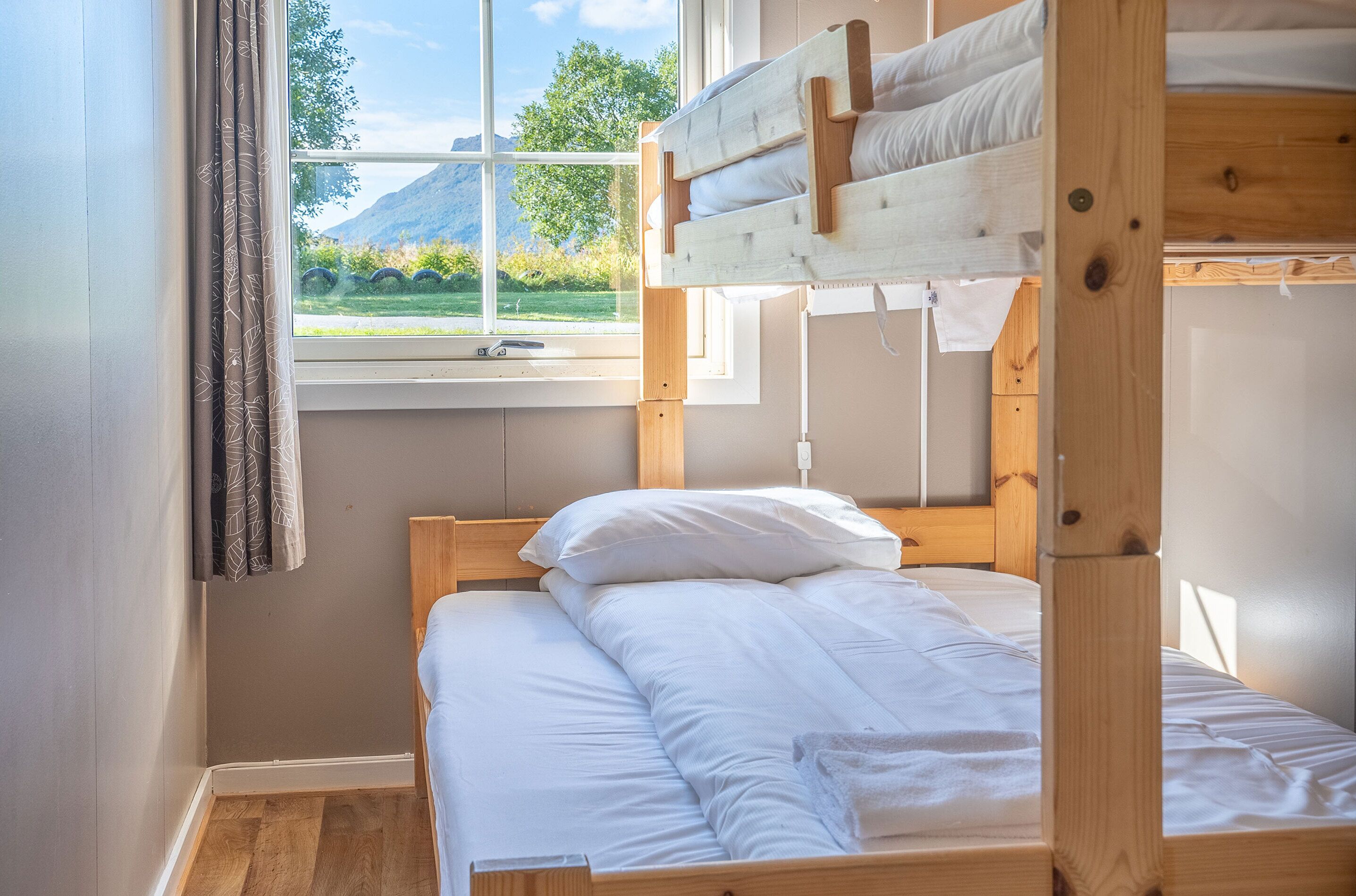 A bunk bed with white bedding, a window with a view of greenery and mountains, and a curtain on the left.