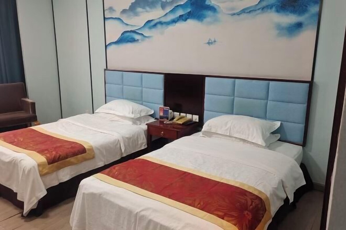 Habitación de hotel con dos camas, cada una con colcha roja y blanca, cabecero azul y un cuadro de montañas en la pared.