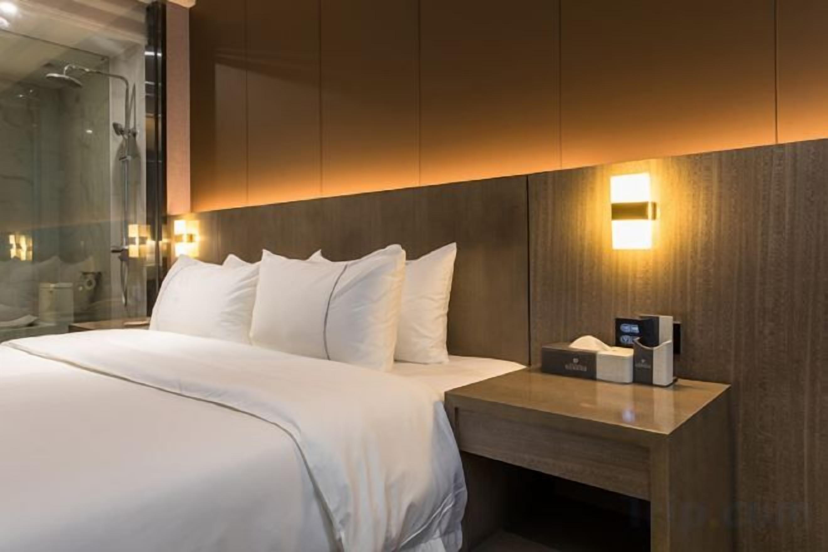Quarto de hotel moderno com uma cama grande, mesa de cabeceira com abajur e box de chuveiro em vidro.