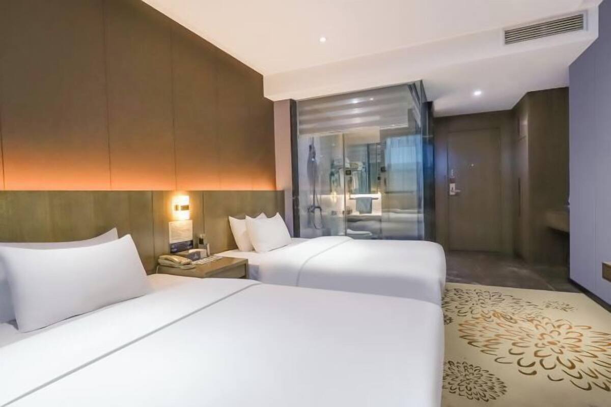 Hotelkamer met twee bedden, een glazen douchecabine, een nachtkastje met lamp en een kamerbreed tapijt met patroon.