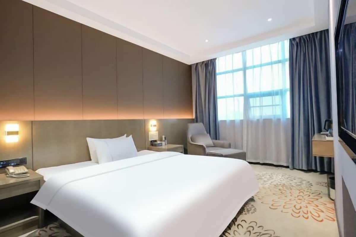 Een hotelkamer met een groot bed, nachtkastjes, een stoel, een bureau en een groot raam met gordijnen.