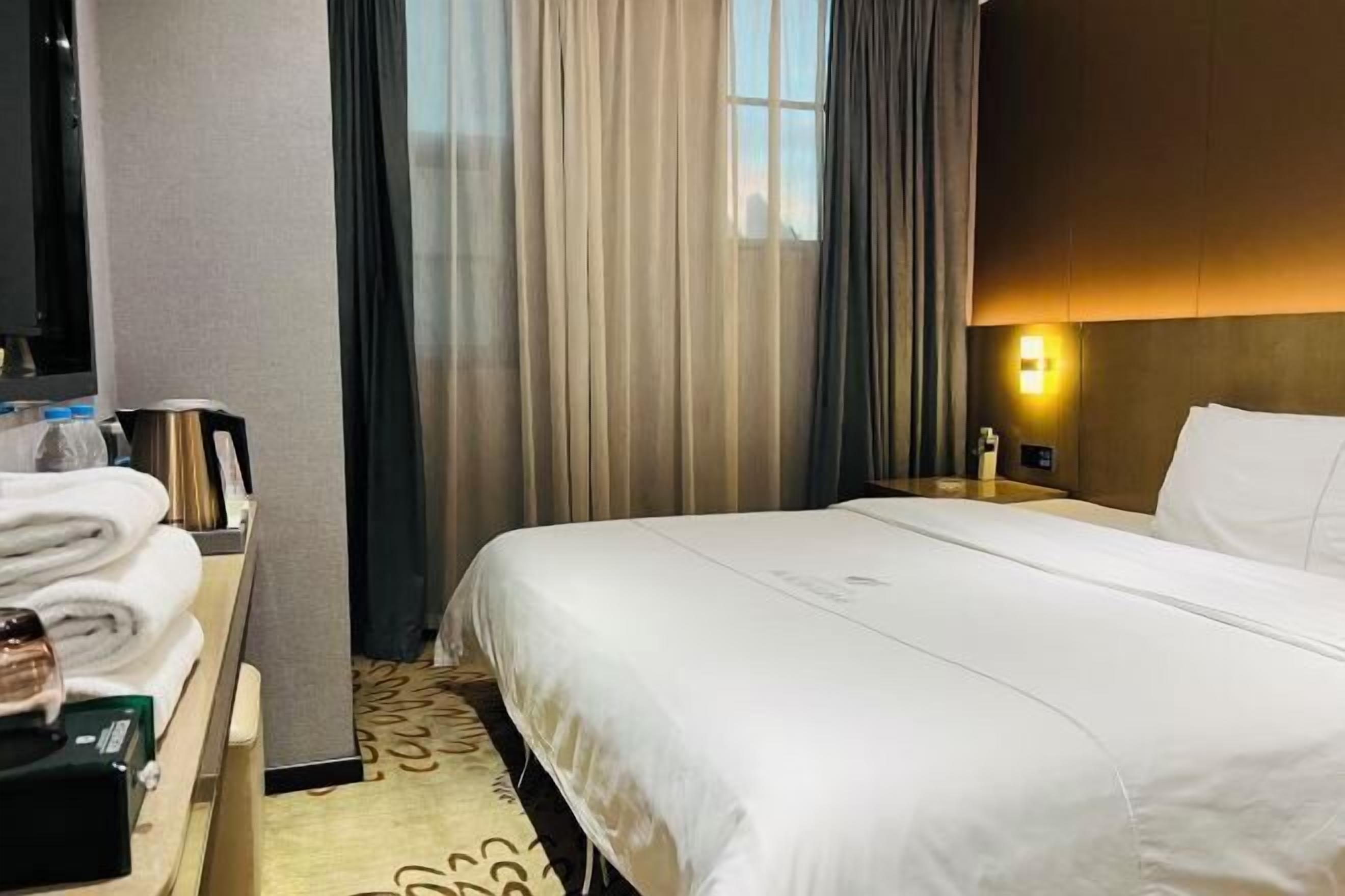 A hotel room with a neatly made bed, a bedside table with a lamp, and a window with curtains.