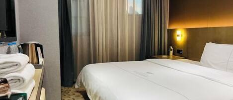 A hotel room with a neatly made bed, a bedside table with a lamp, and a window with curtains.