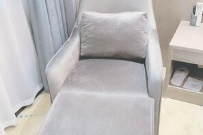 A grey cushioned chair with a footrest, a wooden side table with a remote control, and a pair of slippers on the floor. - Xianglong Boutique Hotel (Guangzhou North Railway Station Huacheng Road Subway Station) (Guangzhou)