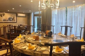 A round dining table set for a meal with chairs, a chandelier, and a view of the outdoors through large windows. - 1881 Peninsula Hotel (Wuxi)