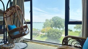 Une pièce dotée d’une grande fenêtre donnant sur un lac et une végétation luxuriante. On y trouve un fauteuil en osier avec un coussin, ainsi qu’une petite table ronde sur laquelle est posé un plateau.