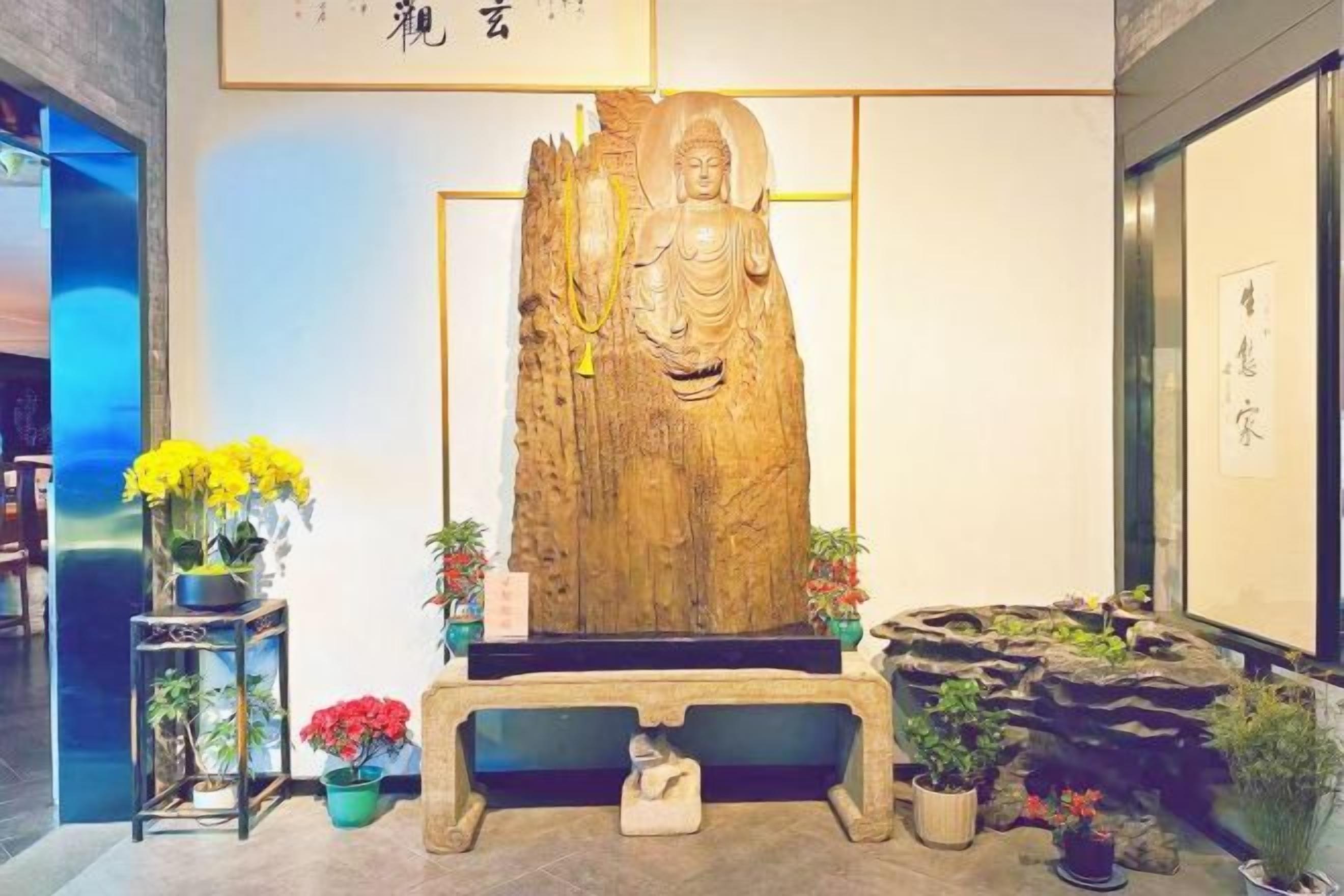 A large wooden statue with a seated figure, surrounded by potted plants and a stone bench.