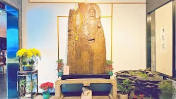 Een groot houten beeld met een zittende figuur, omringd door potplanten en een stenen bank.