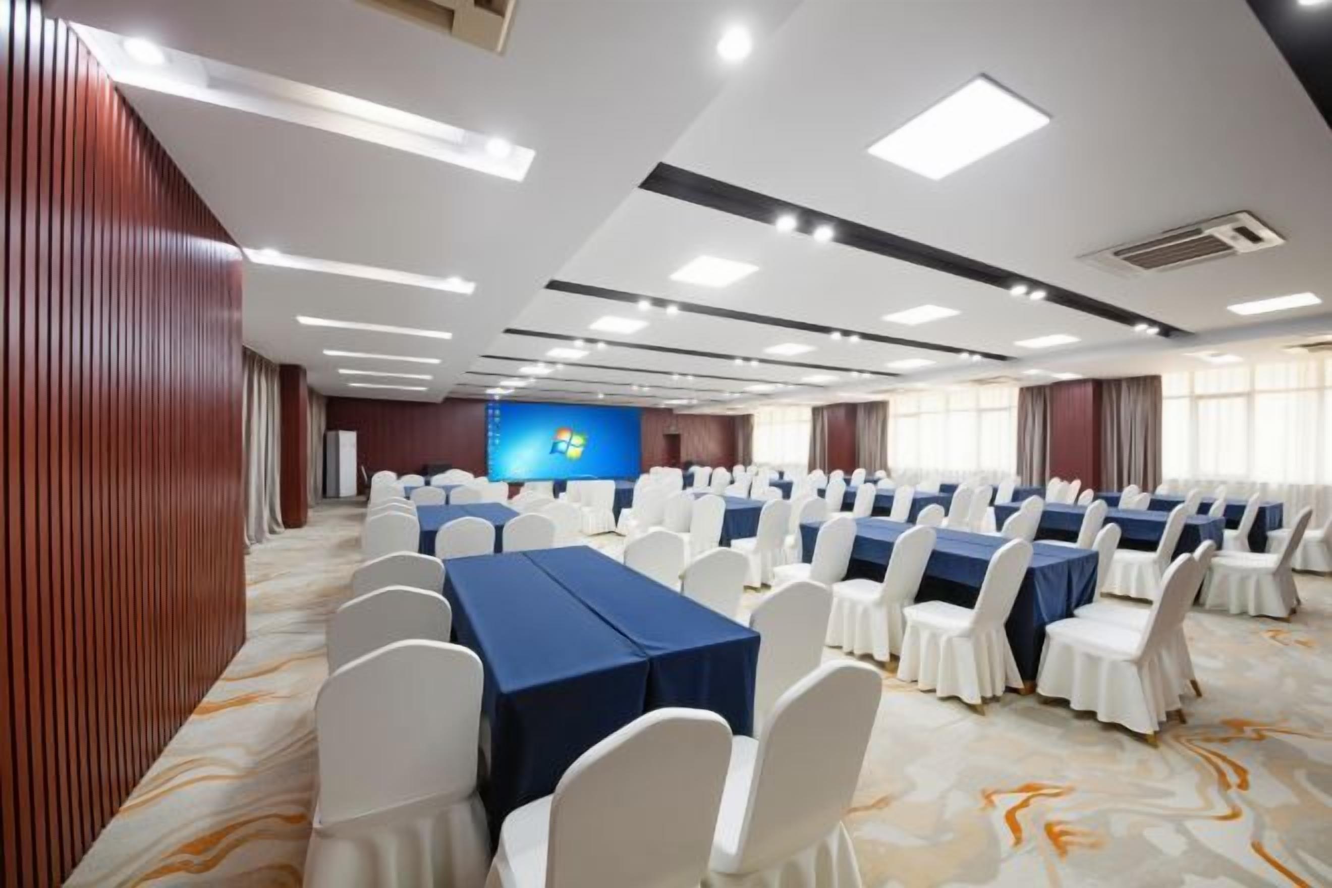 Un salón de conferencias con filas de mesas y sillas, una pantalla grande y suelo alfombrado.