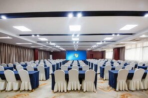 Un salón de conferencias con filas de mesas y sillas, un escenario con una pantalla y suelo alfombrado.