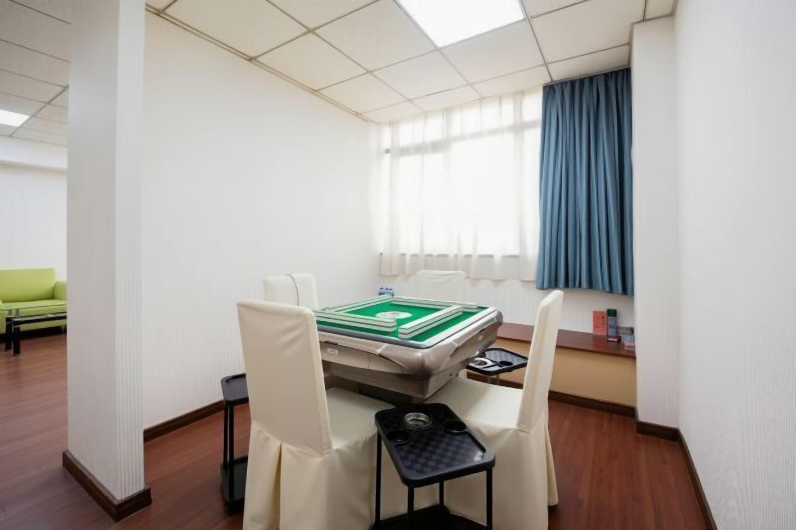 Habitación de hotel con cama, una mesita, dos sillas, ventana con cortinas y un sofá verde en la habitación contigua.