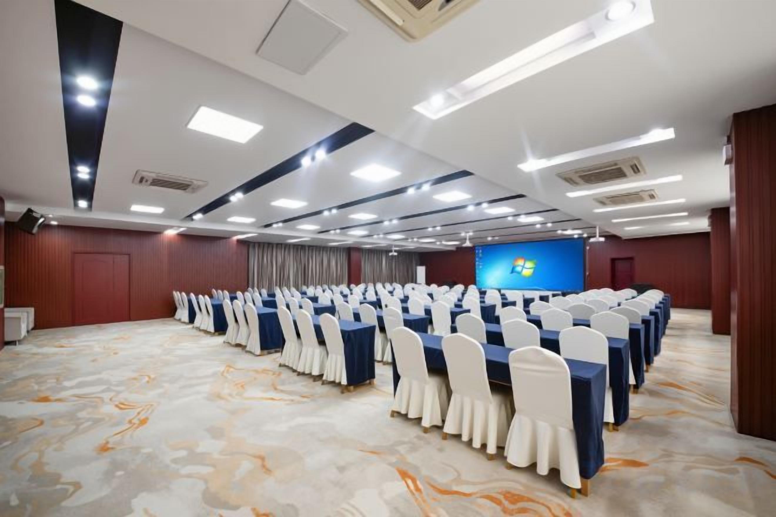 Egy konferenciaterem, melyben sorokban állnak az asztalok és székek, egy nagy képernyő és szőnyeg borítja a padlót.