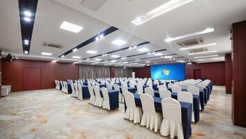 Un salón de conferencias con filas de mesas y sillas, una pantalla grande y piso alfombrado.