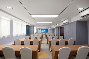Un salón de conferencias con filas de mesas y sillas, una pantalla grande al frente y una ventana a la izquierda.
