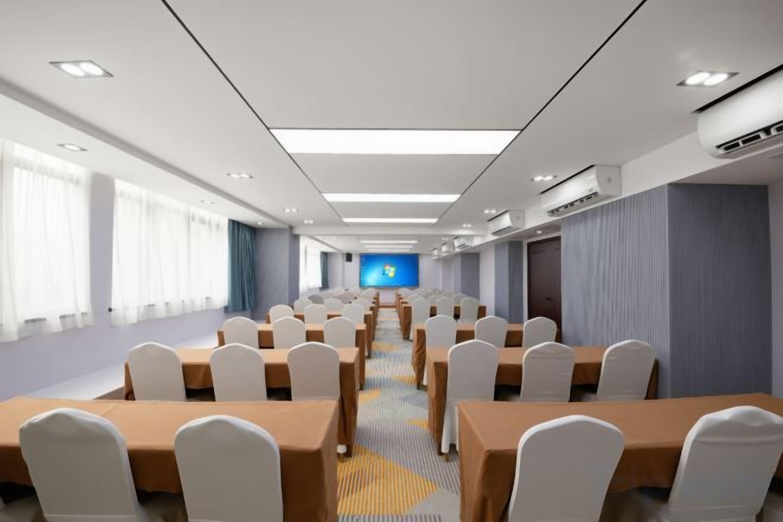 Un salón de conferencias con filas de mesas y sillas, una pantalla grande al frente y una ventana con cortinas.