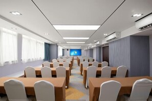 Un salón de conferencias con filas de mesas y sillas, una pantalla grande al frente y una ventana con cortinas.