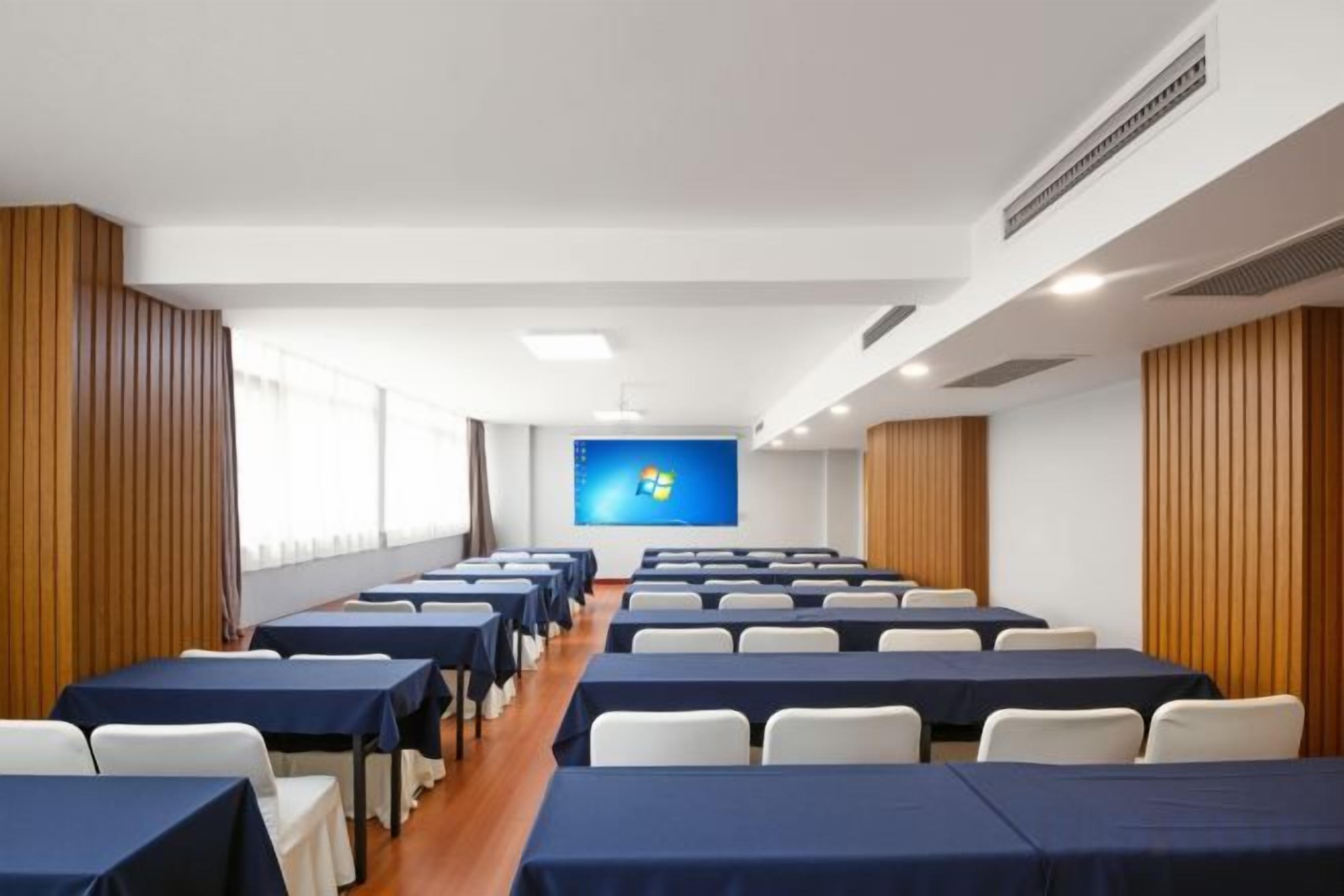 Un salón de conferencias con filas de mesas y sillas, una pantalla grande al frente y paneles de madera en las paredes.