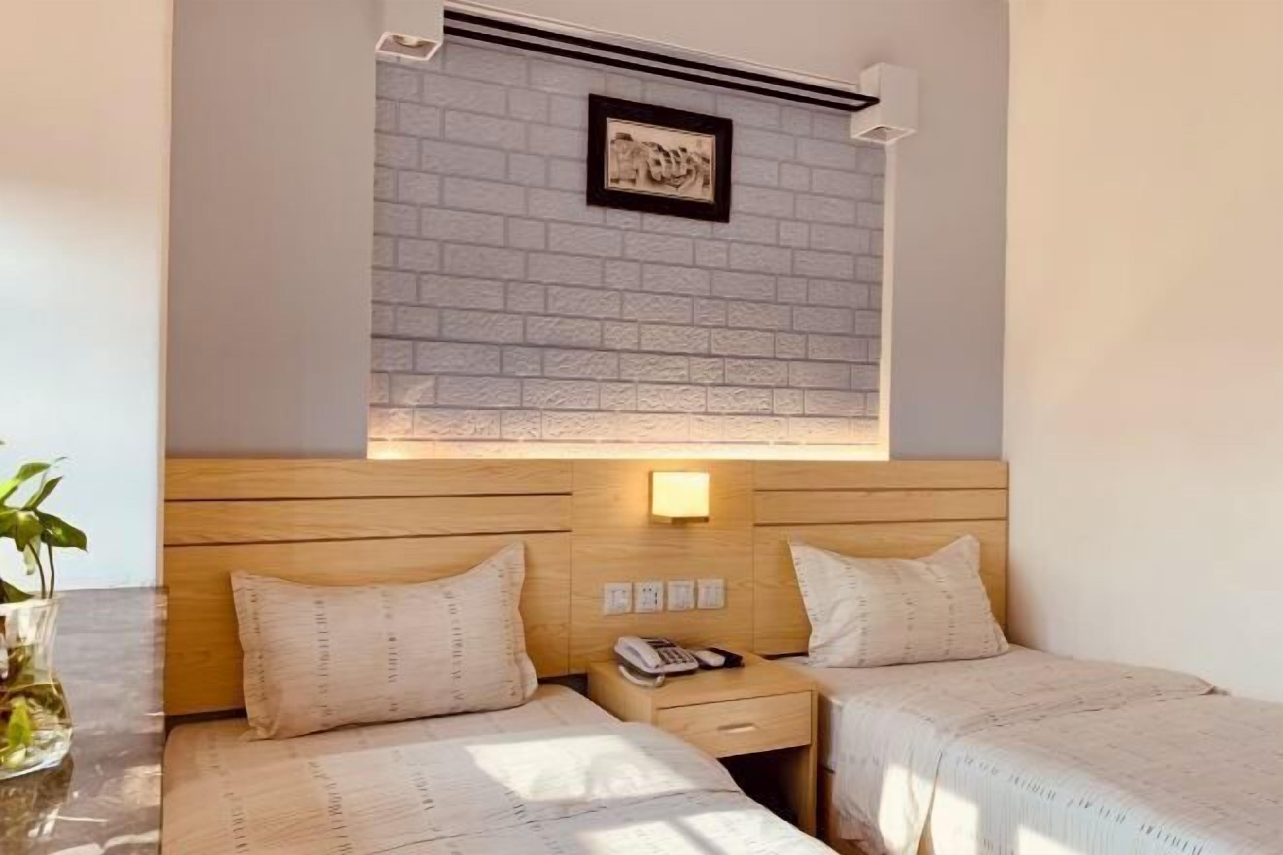 Habitación de hotel con dos camas, cabecero de madera, una mesita con un florero y una lámpara fijada a la pared.