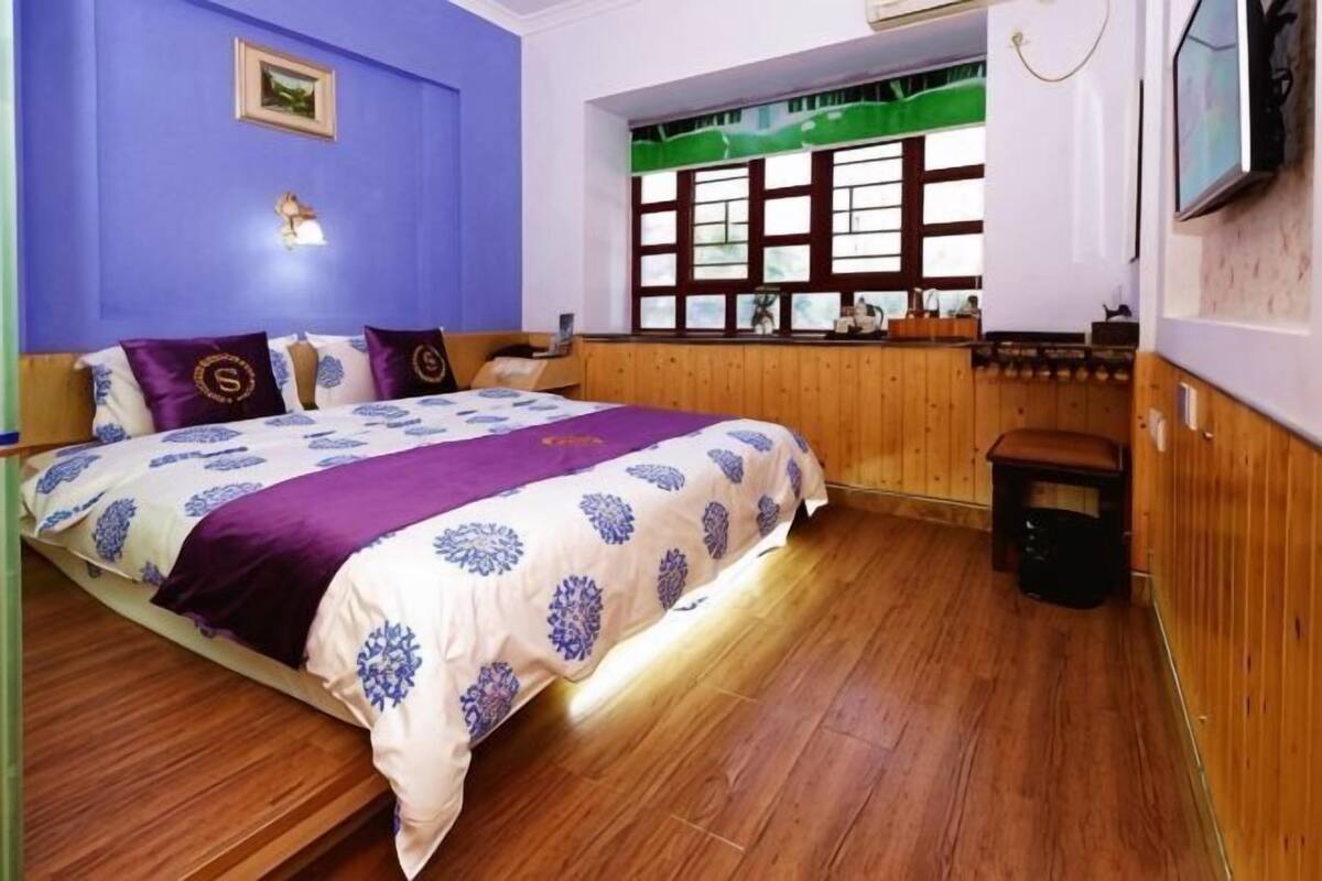 A bedroom with a bed, wooden flooring, a window with blinds, a wall-mounted TV, and a small stool.