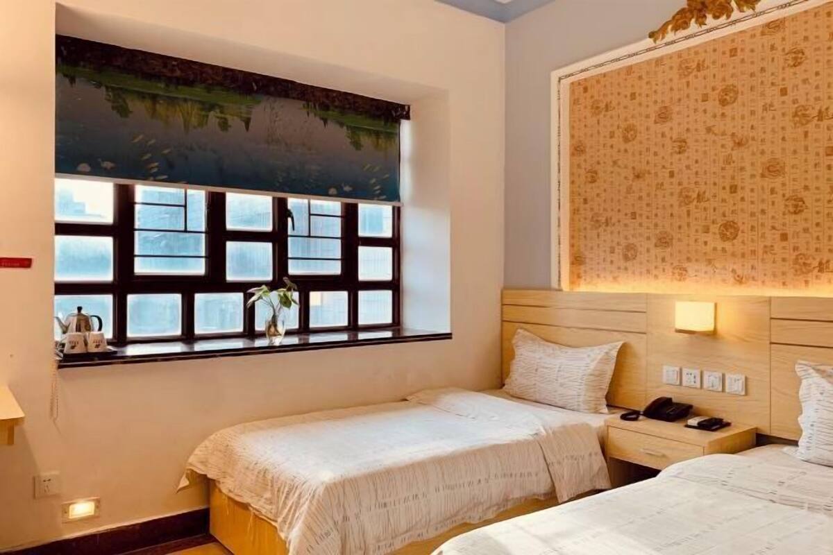 A hotel room with two beds, a window with a view, a wall-mounted lamp, and a nightstand with a phone and a kettle.