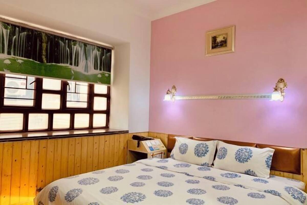 A bedroom with a bed, wooden paneling, a window with a view, a wall-mounted shelf, and a picture.
