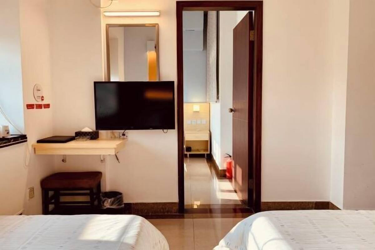 A hotel room with a bed, a desk with a flat-screen TV, a small stool, and a mirror on the wall.