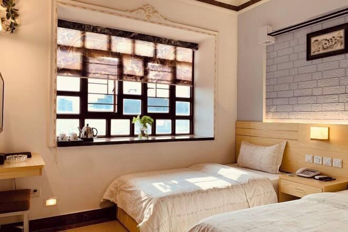 A hotel room with two beds, a window with a view, a bedside table with a lamp, and a wall-mounted phone.