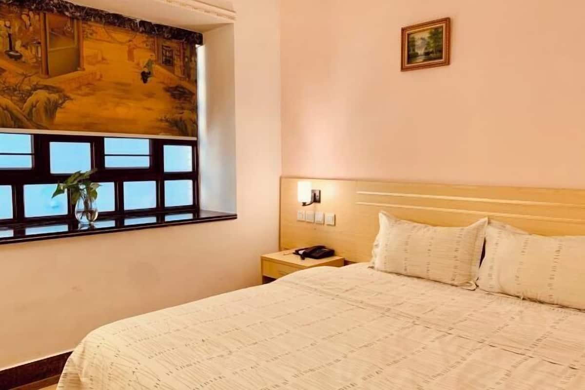 A hotel room with a large bed, a nightstand with a lamp, a window with a view, and a painting on the wall.
