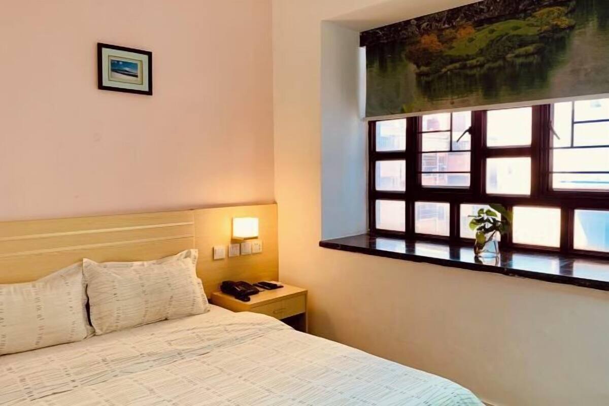 A hotel room with a bed, bedside table, lamp, and a window with a view of a landscape.