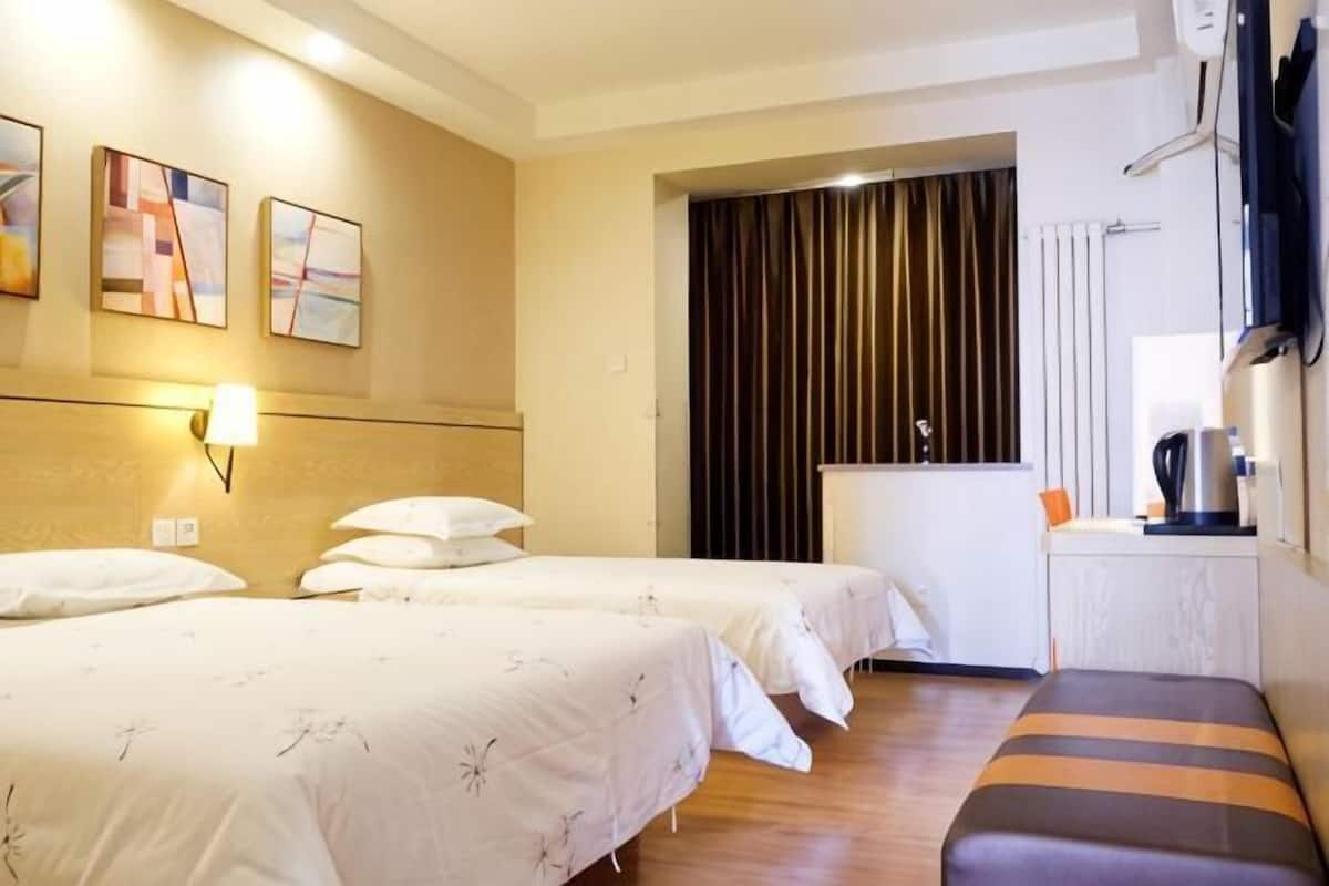 A hotel room with two beds, a bench, a desk with a kettle, and a window with curtains.