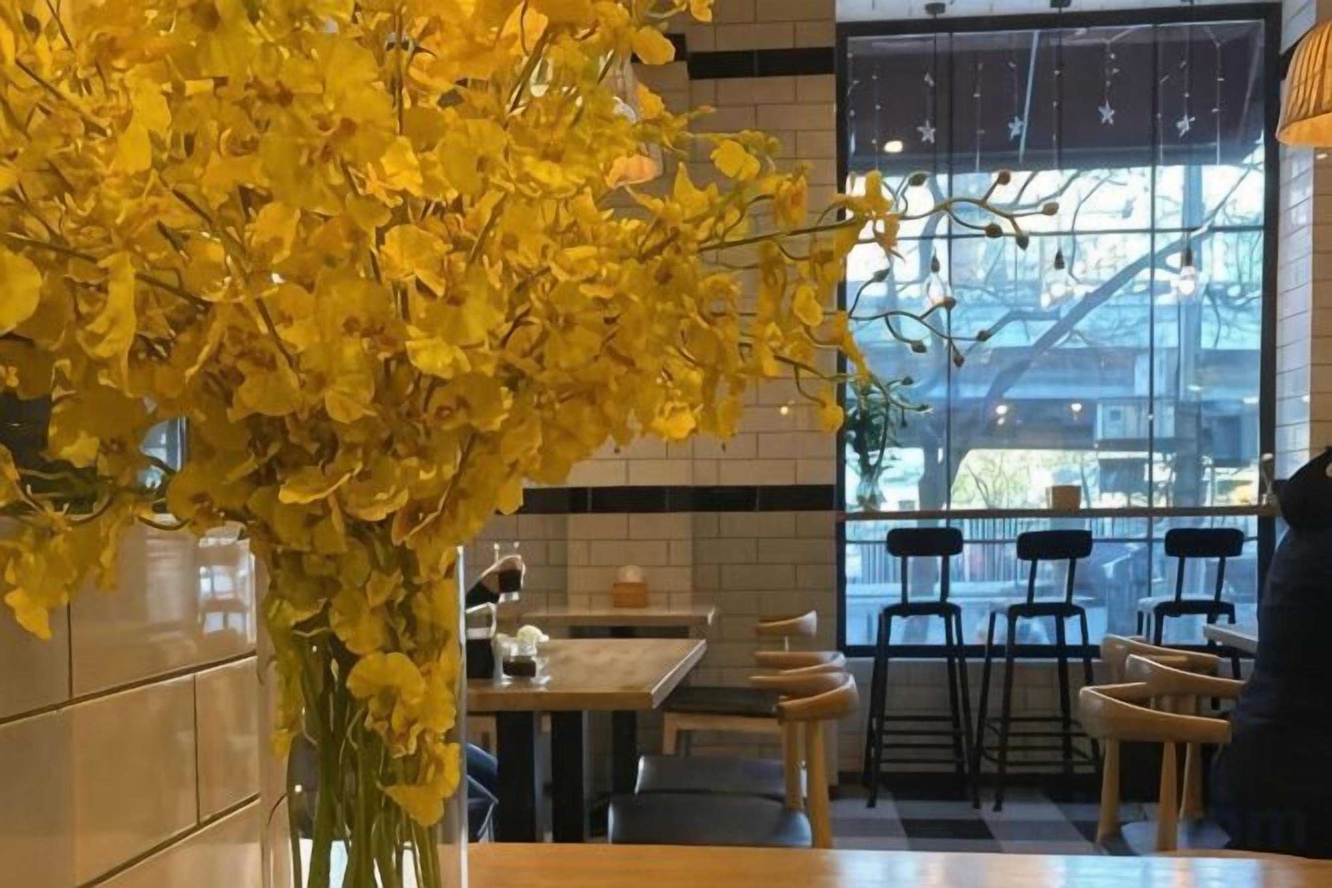 Interior de un restaurante con un ventanal grande, mesas, sillas y un jarrón con flores amarillas.
