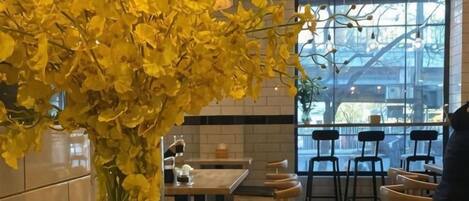 A restaurant interior with a large window, tables, chairs, and a vase of yellow flowers.