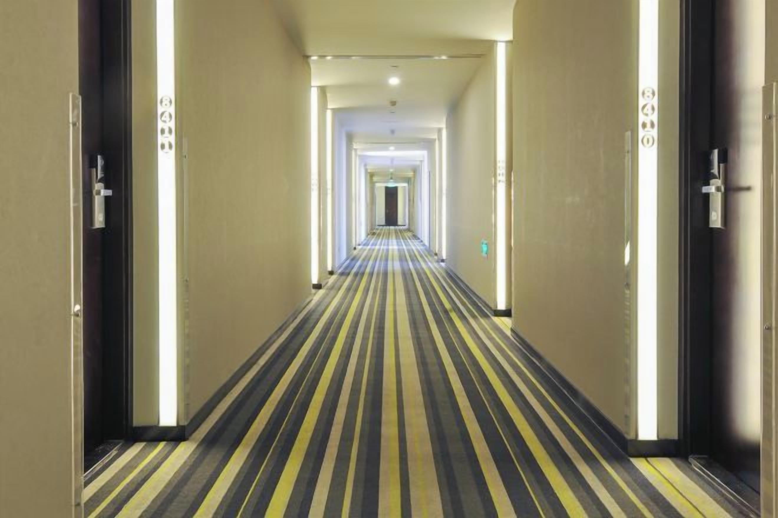 Een hotelgangen met een gestreept tapijt, deuren aan beide kanten, en een lange gang die uitkomt in een helder verlichte ruimte.