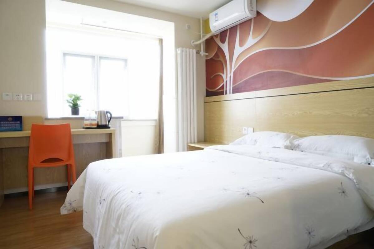A hotel room with a bed, a desk, an orange chair, a window, and a wall with a floral mural.