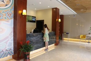 Zona de recepción de hotel con un mostrador de mármol, una pantalla digital y un área de descanso con una silla amarilla.