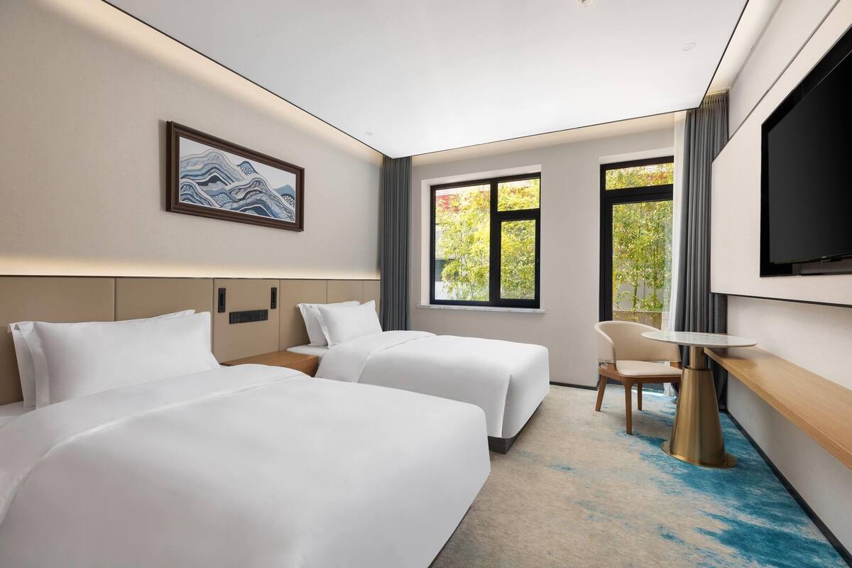 A hotel room with two beds, a desk, a chair, a TV, and a window with a view of trees.