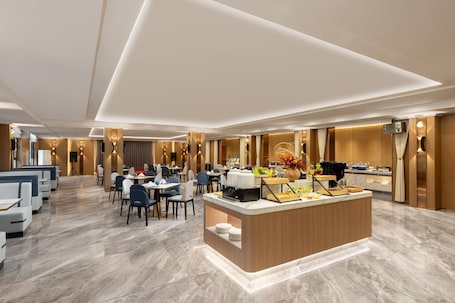 A modern hotel dining area with a central buffet station, multiple seating arrangements, and a polished marble floor.