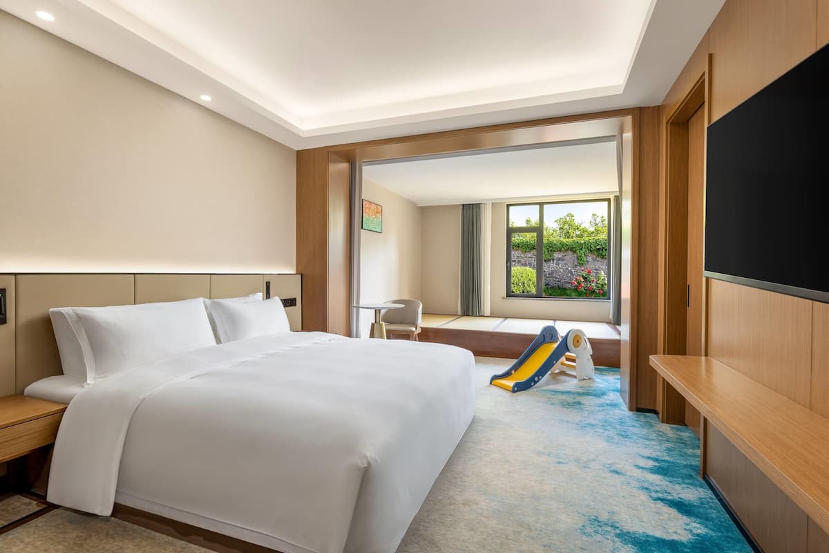 A hotel room with a large bed, a desk, a TV, and a window with a view of greenery.