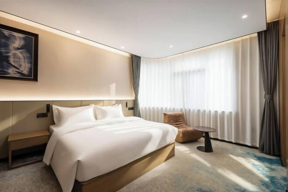 A modern hotel room with a large bed, a small side table, a chair, and a large window with curtains.