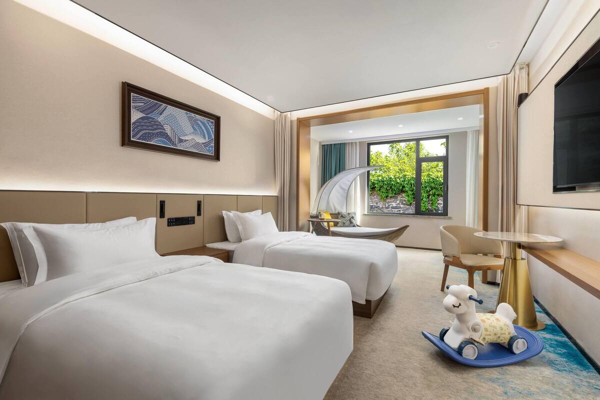 A hotel room with two beds, a desk, a chair, a TV, and a window with a view of greenery.