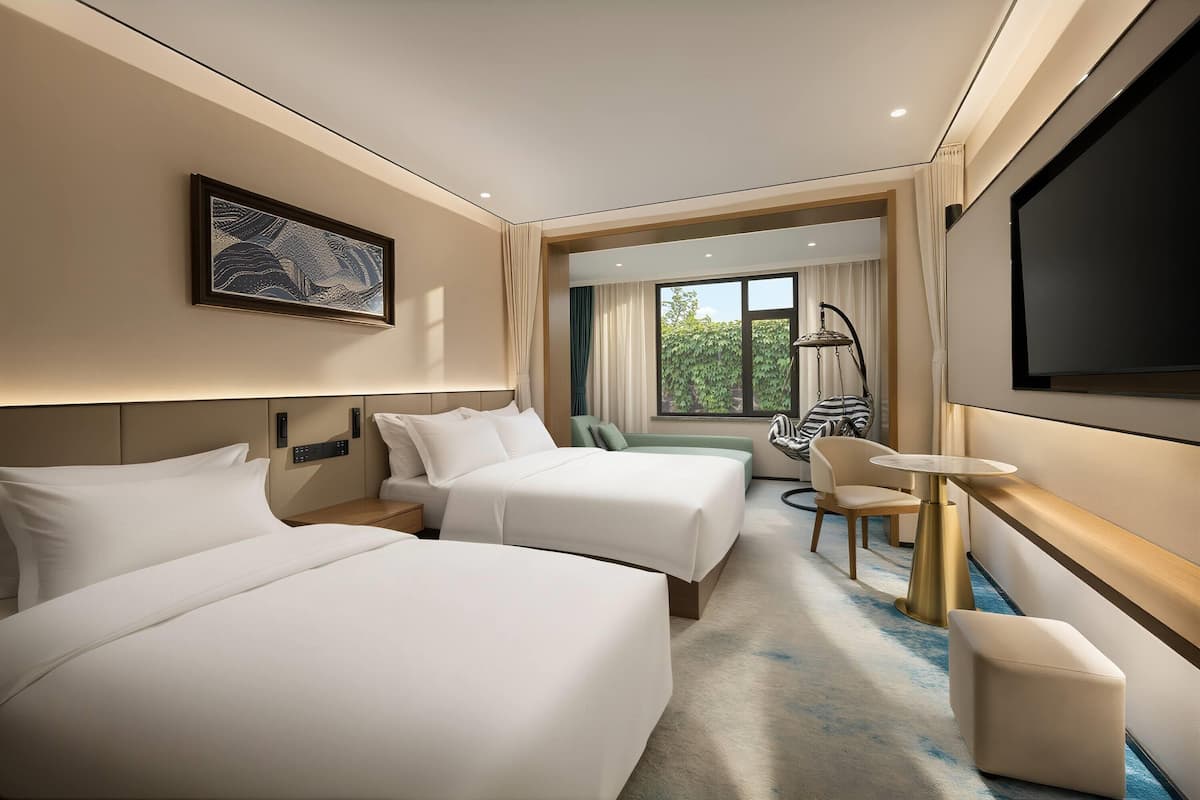 A hotel room with two beds, a TV, a desk, and a view of greenery.