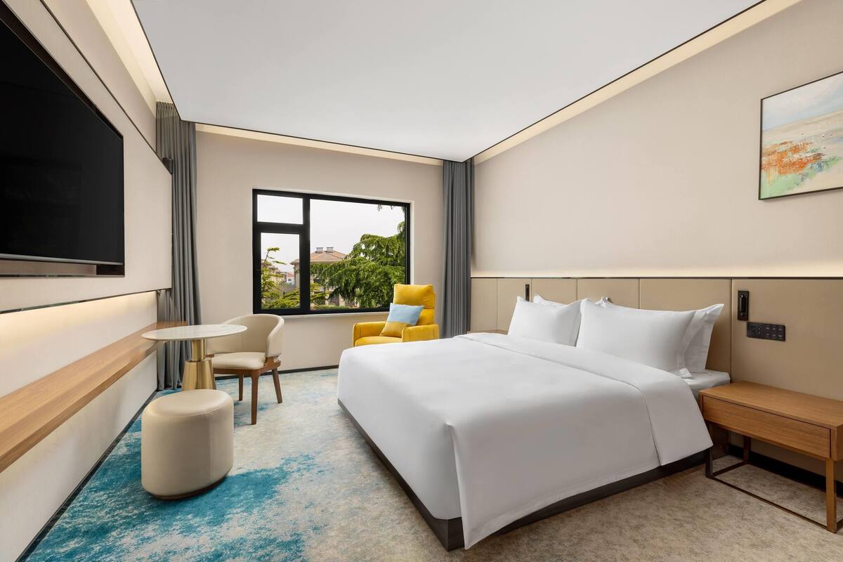A modern hotel room with a large bed, a desk, a chair, a TV, and a window with a view.