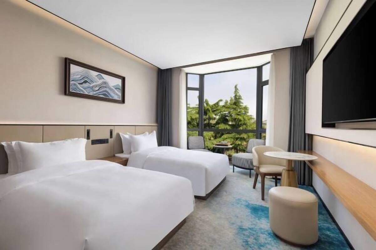 A hotel room with two beds, a small table, a chair, a TV, and a large window with a view of greenery.