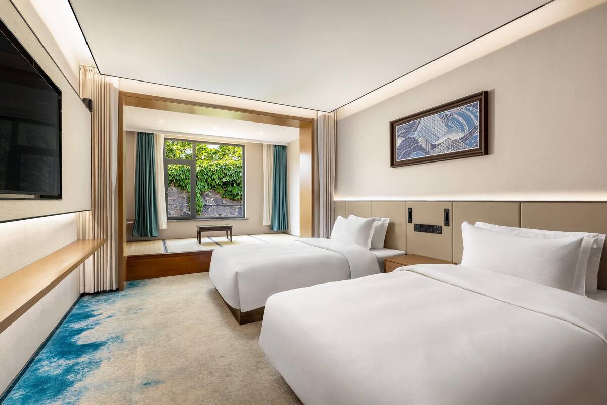 A hotel room with two beds, a large window with curtains, a flat-screen TV, and a framed artwork on the wall.