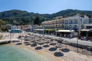 Ein Küstenhotel mit mehreren Balkonen, ein Strandbereich mit Sonnenschirmen und eine Bergkulisse im Hintergrund.