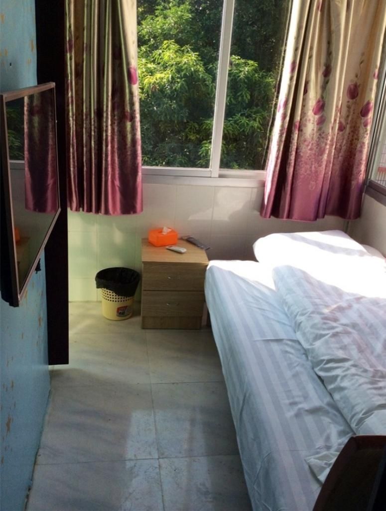A small room with a bed, a nightstand, a window with curtains, and a wall-mounted shelf.