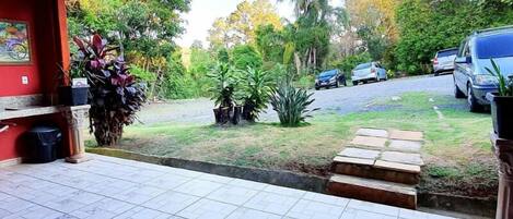 A covered outdoor area with tiled flooring, a bench, potted plants, and a view of a road with parked cars.