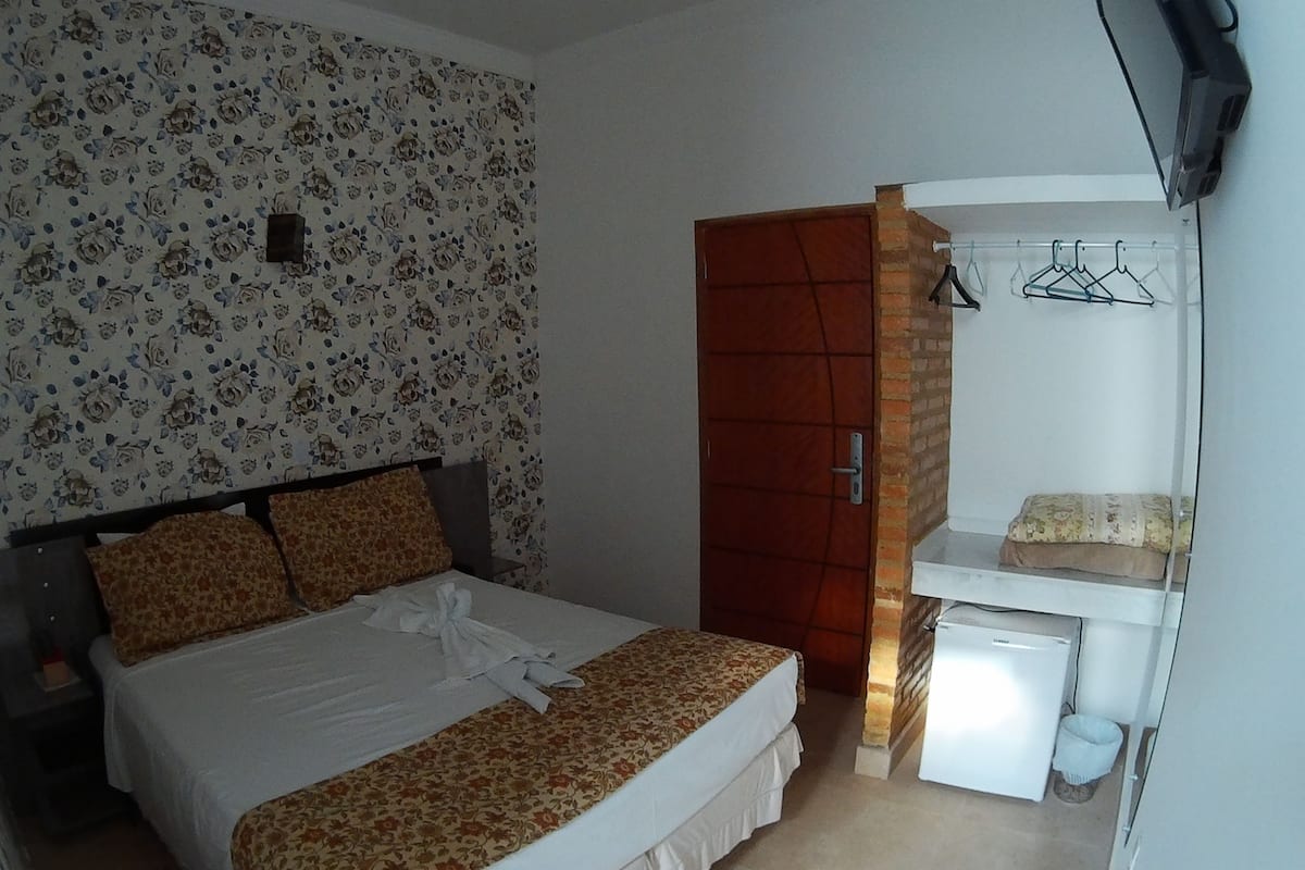 Un dormitorio con cama, armario, refrigerador blanco pequeño y un espejo con ropa colgada.