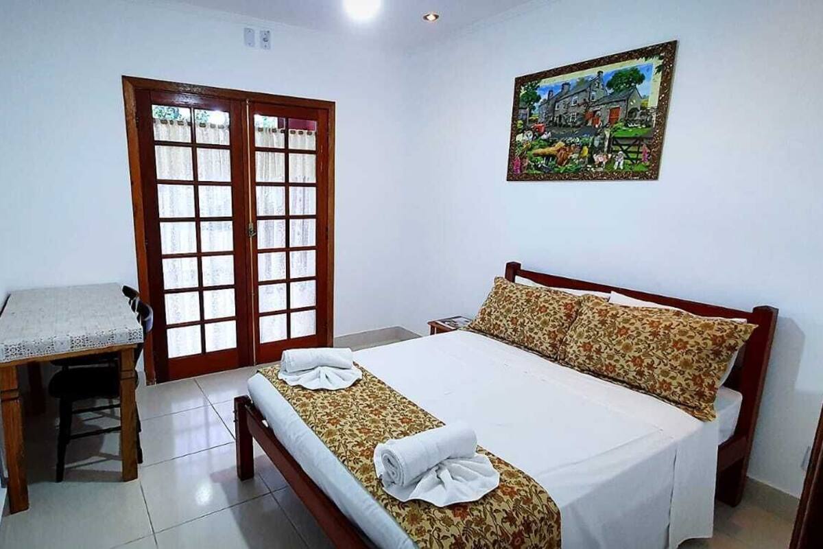 Un dormitorio con cama, una mesita, una silla, una puerta con paneles de vidrio y un cuadro en la pared.